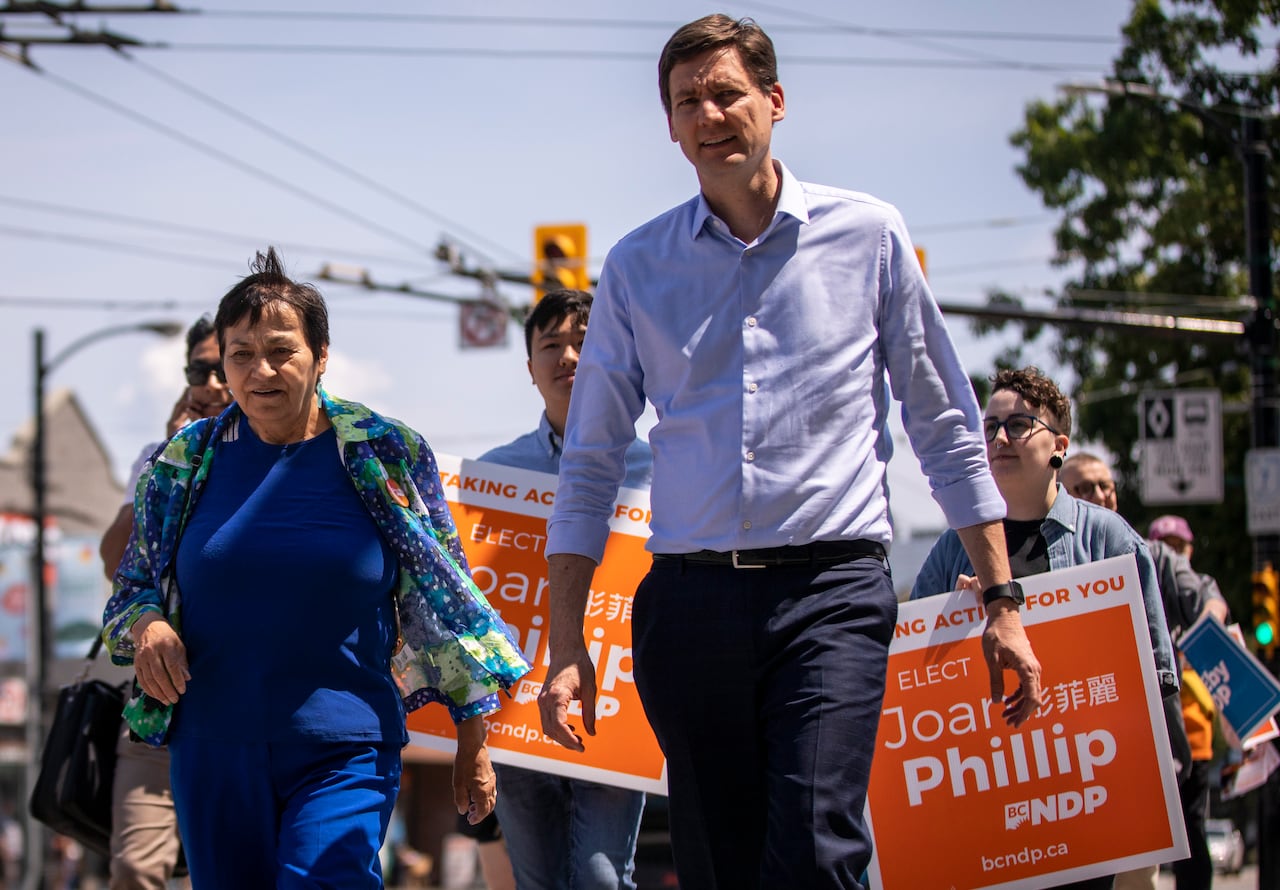 A tall white man is seen next to an Indigenous woman, with people behind them carrying orange signs.