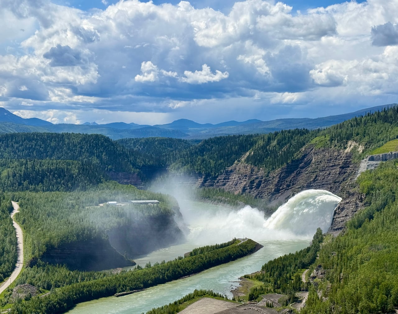 a giant misty waterfall from surges from a dam spillway through a forested mountain canyon and into a winding river below