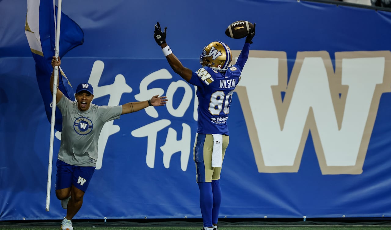 A football player wearing a blue and gold jersey with the number 80 and last name "Wilson" raises his hands in the air and celebrates with a ball in his right hand. A person wearing a grey shirt waves a flag and celebrates