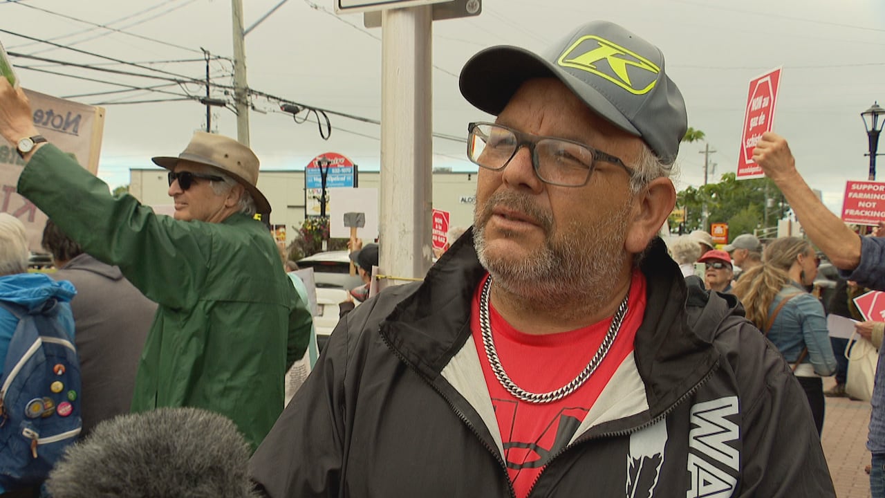 A man being interviewed with people holding protest signs behind him.
