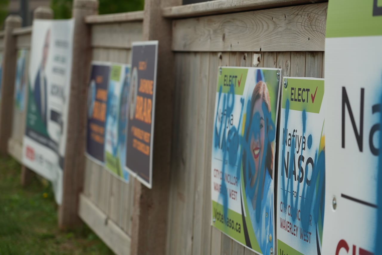 A variety of signs along a fence, some of them belonging to Naso, are covered with markings in blue spray paint.