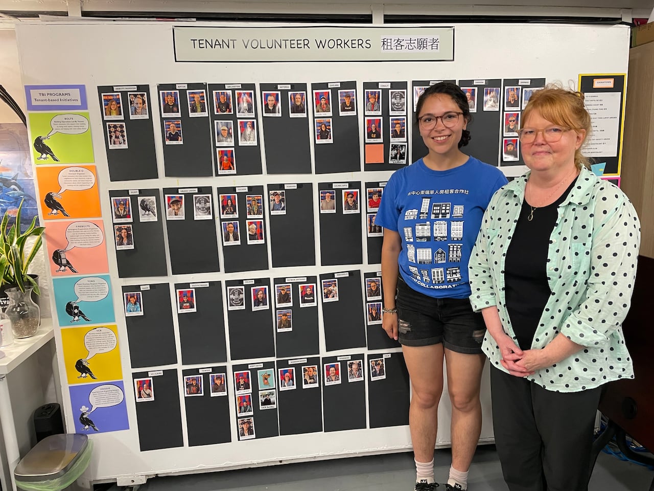 Two women pose next to a display board with photos of people and their names.