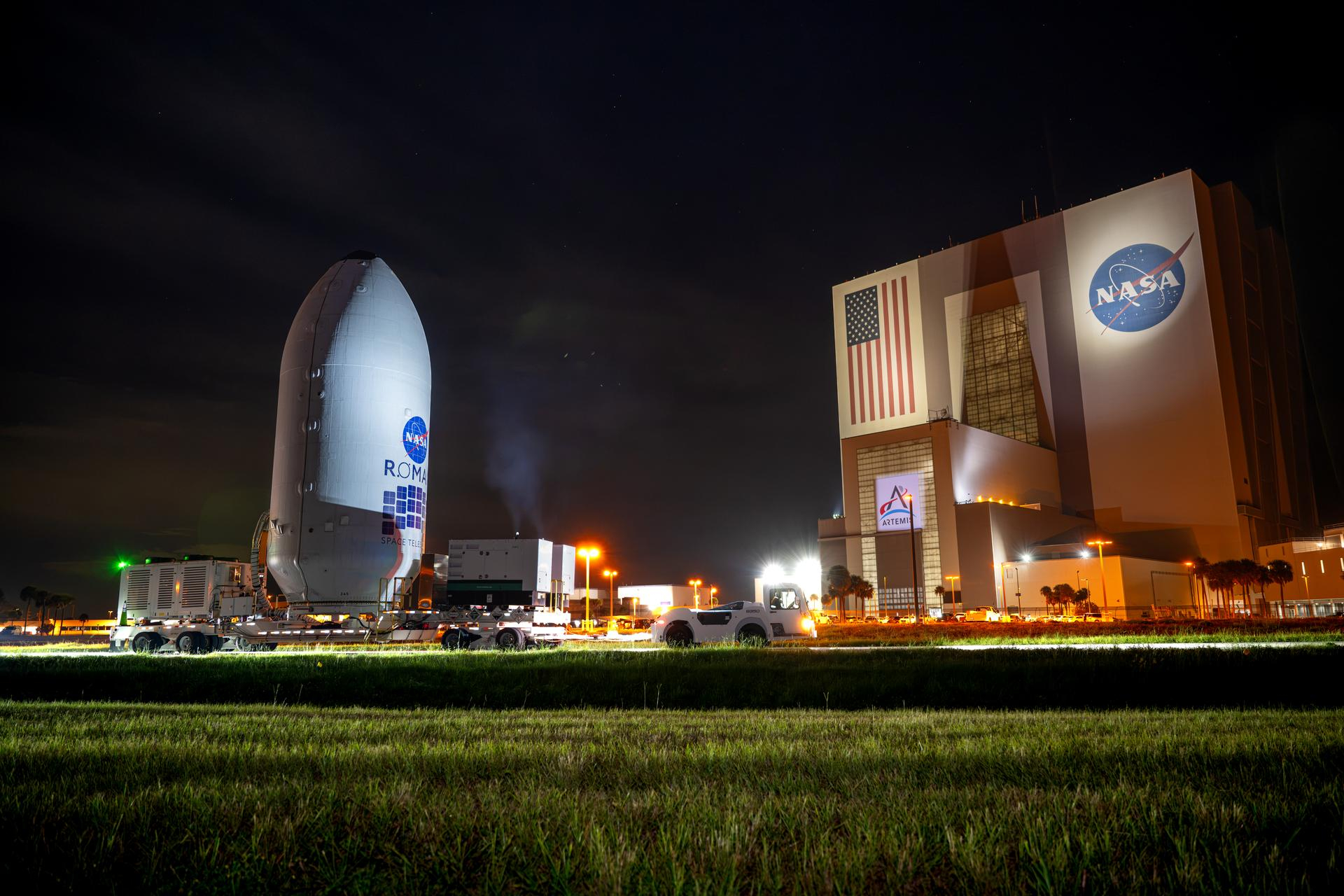 A white, rounded point cylindrical capsule is rolled by a giant cube building.
