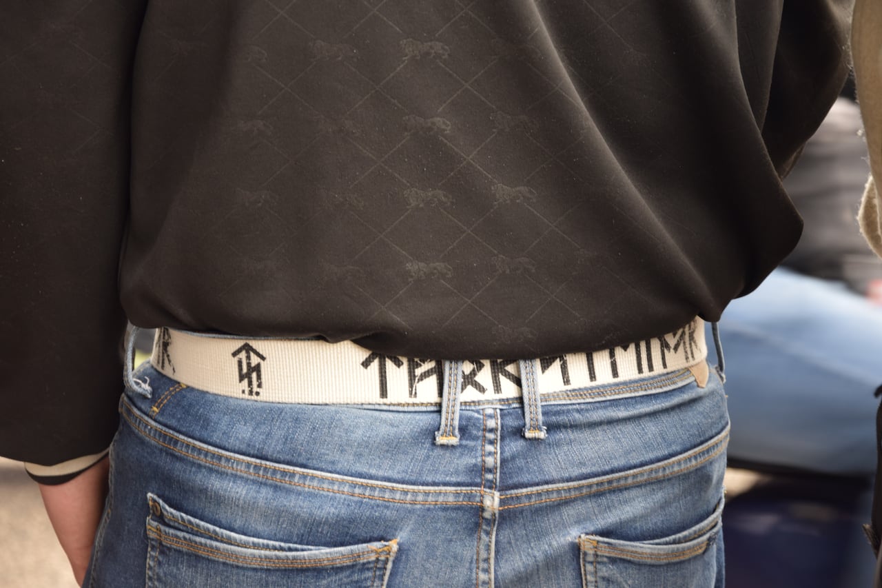 A man in a black t-shirt is seen wearing a nylon belt in black with white Nordic rune symbols on it.