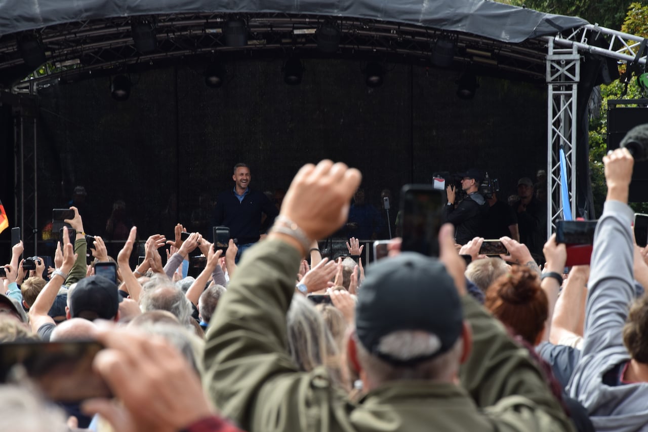 A group of people is seen cheering towards a stage with a political leader giving a speech