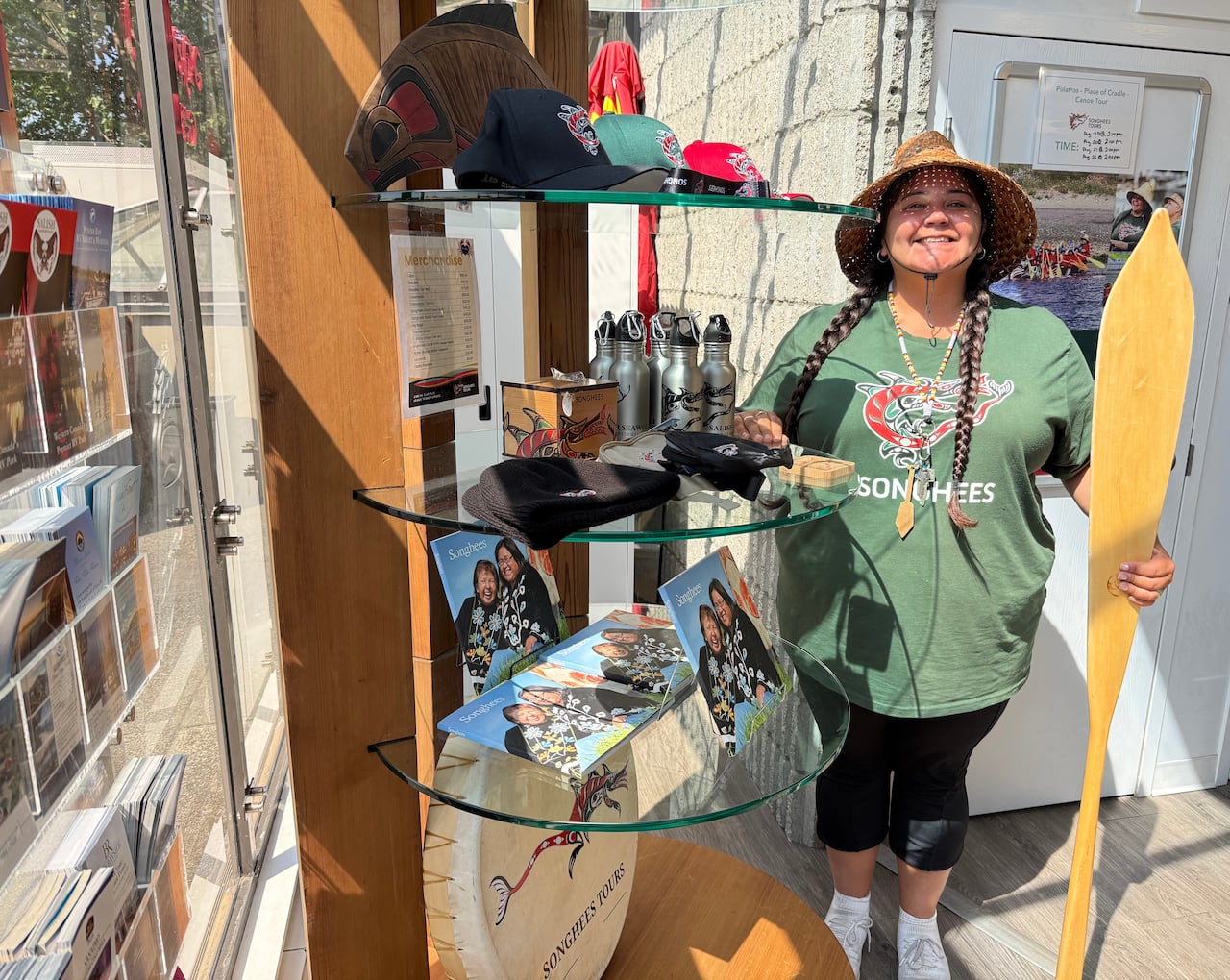 An Indigenous woman holds an oar next to a rack with pictures and merchandise on it.