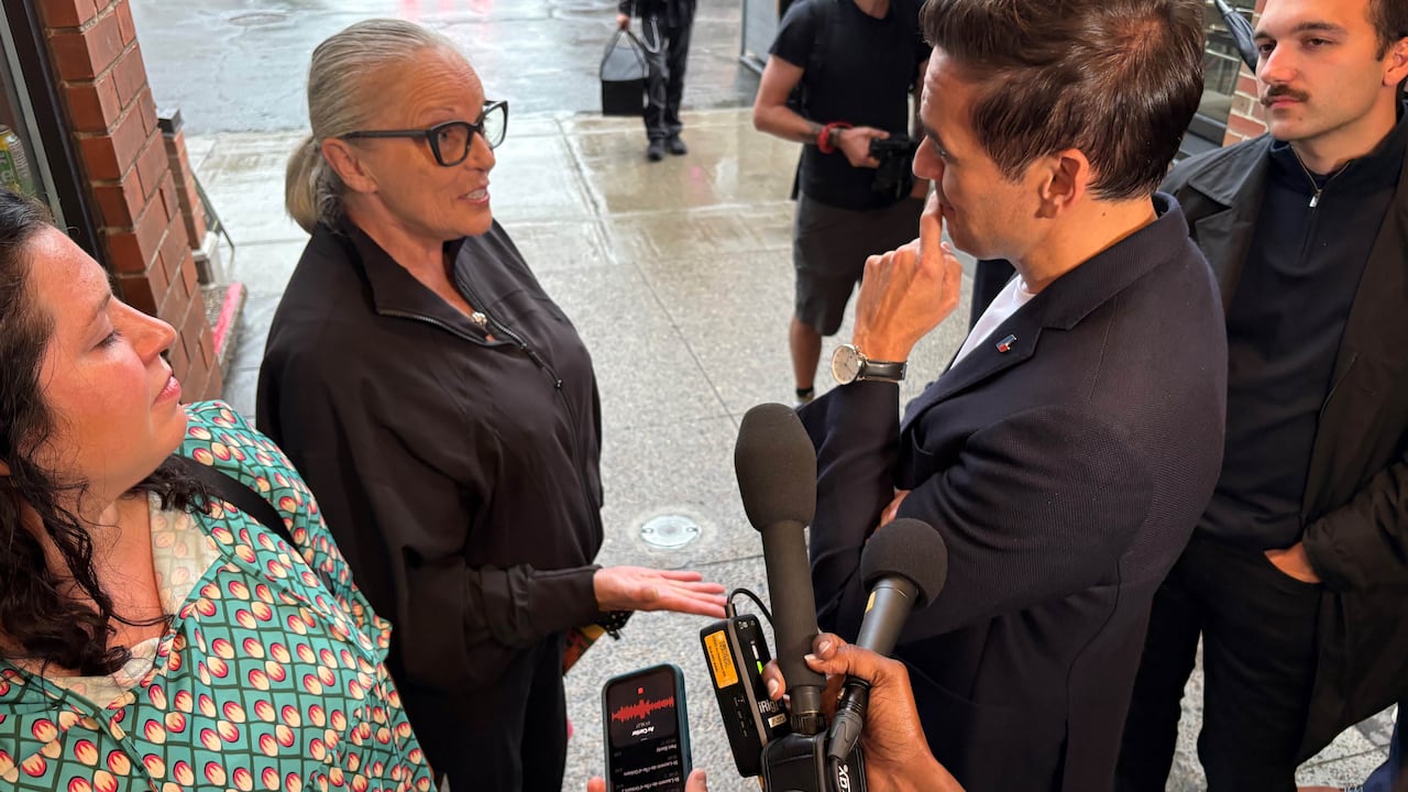 An elderly woman speaks to a politician while reporters record the interaction.