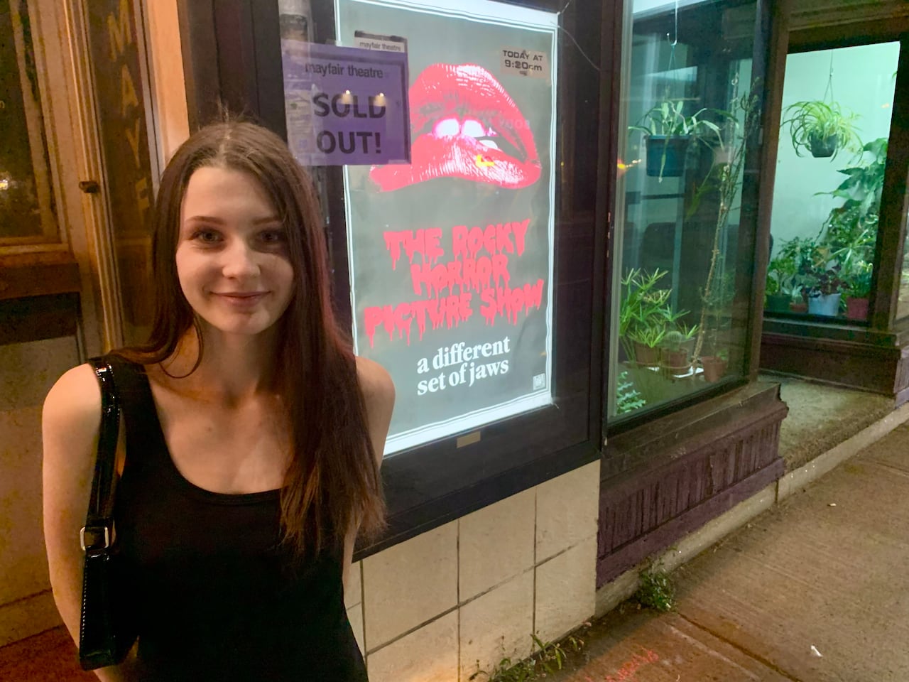 A woman in a black dress stands by a movie poster.