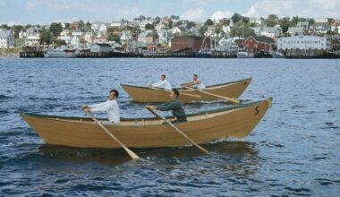 Pairs of white male rowers in two red open boats called dories pull their oars through the water with a seaside town in the background.