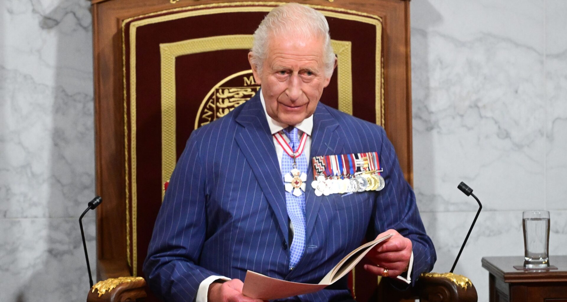 King Charles on a throne-like chair, wearing medals and holding an open book.