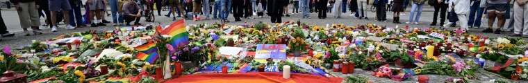 A large rainbow flag is seen on the pavement in front of the Brandenburg Gate, with flowers and other items near it as onlookers stand nearby.