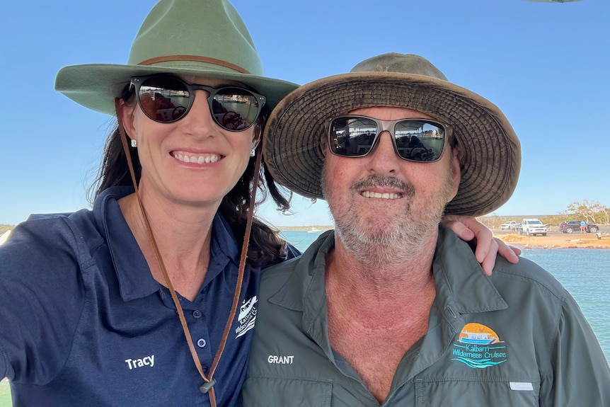 A man and a woman smiling taking a selfie, both in wide brimmed hats and sunglasses, blue water in background.