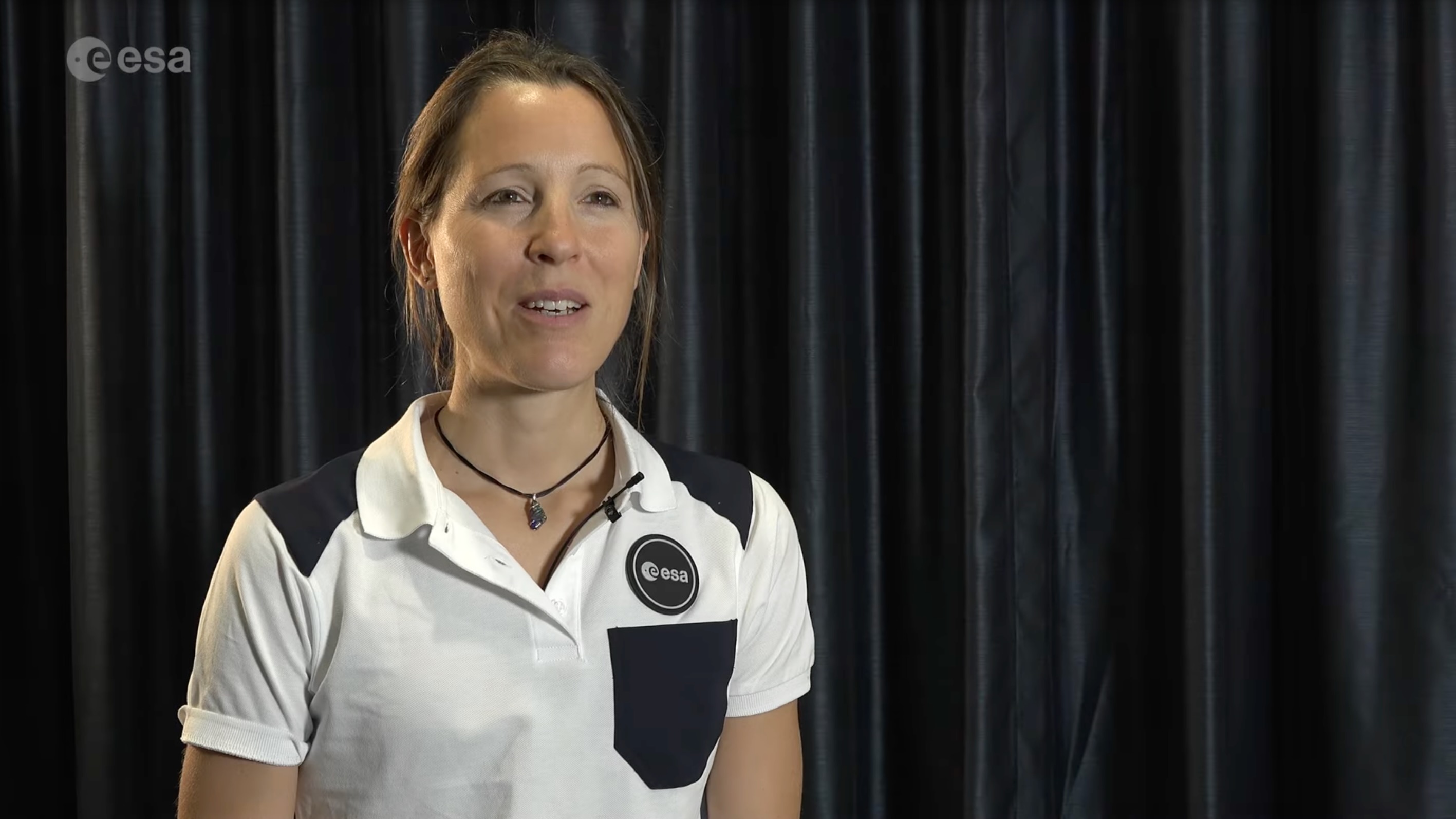 a smiling woman in a white polo shirt sits in front of black drapes