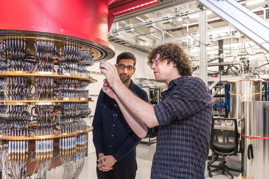 Two men stand next to a vertical cylinder array of wires and chips in a white computer lab setting.