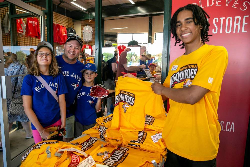 BROOK JONES / FREE PRESS
The Winnipeg Goldeyes take on a pesky and all-too-familiar alternate identity, the Manitoba Mosquitoes, for the team’s home game against the visiting Lincoln Saltdogs on Friday. Kataali Stoller (right) shows off the special edition merchandise to Cory Ruppanthal and his children, Ava and Owen.