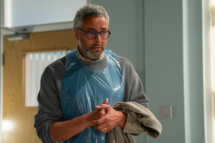 Christopher Colquhoun as Darnell Larson Jr in EastEnders. Darnell, wearing a plastic blue apron, stands in a hospital in a scene from EastEnders