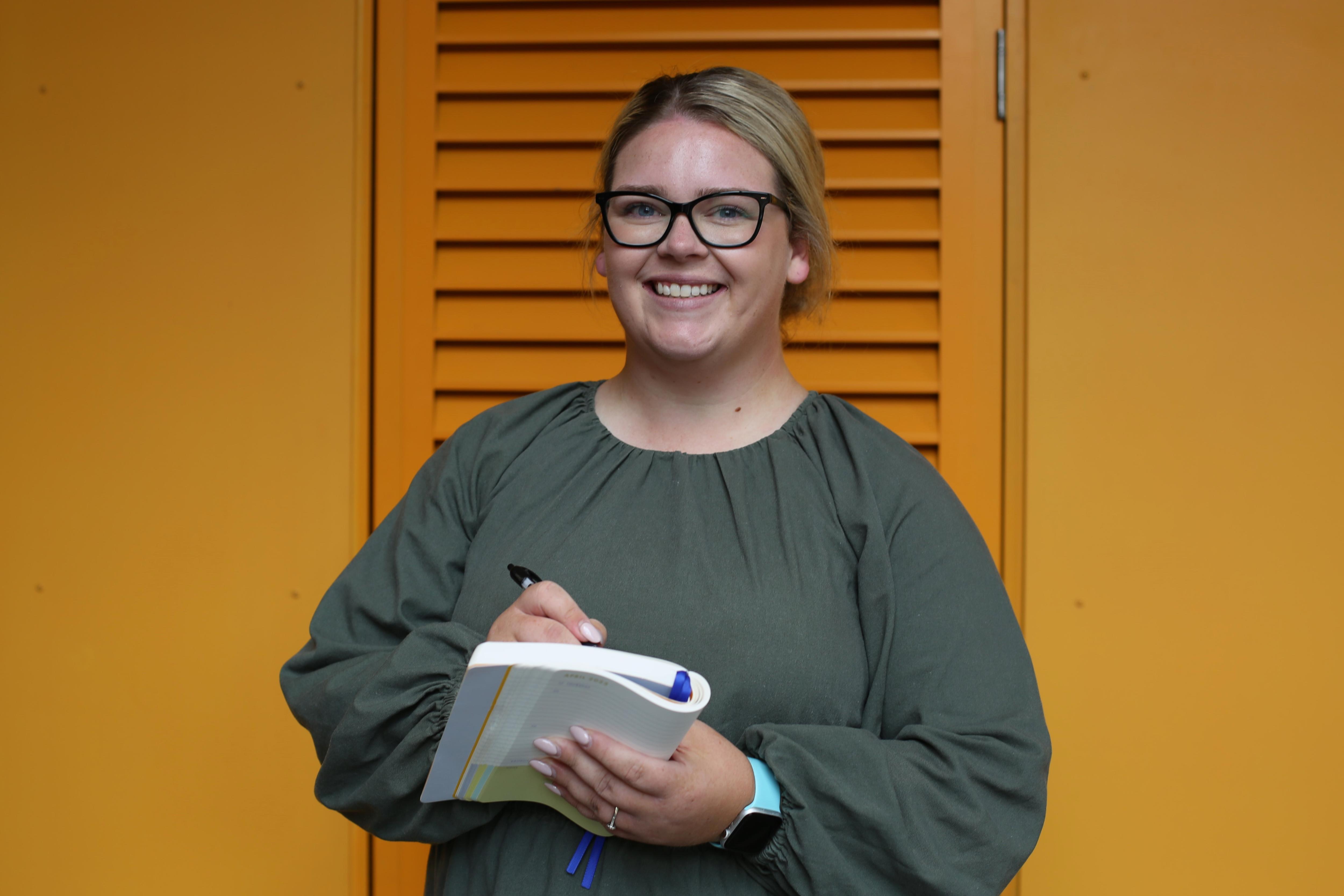 A photo of Alyssia with her blonde hair in a bun, wearing black glasses. She's holding a notebook and pen.