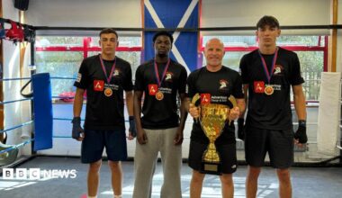 Winners from Odyssey Boxing Club pictured with Ross Getty, who is holding a gold trophy. Two boys to the left of Ross, and one to the right, are all wearing medals around their necks. All four are wearing black t-shirts for the club, with all but one wearing black shorts. They are all standing in the boxing ring.