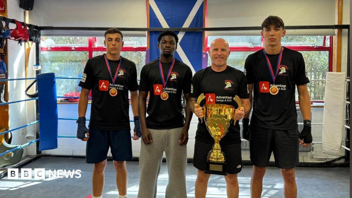 Winners from Odyssey Boxing Club pictured with Ross Getty, who is holding a gold trophy. Two boys to the left of Ross, and one to the right, are all wearing medals around their necks. All four are wearing black t-shirts for the club, with all but one wearing black shorts. They are all standing in the boxing ring.