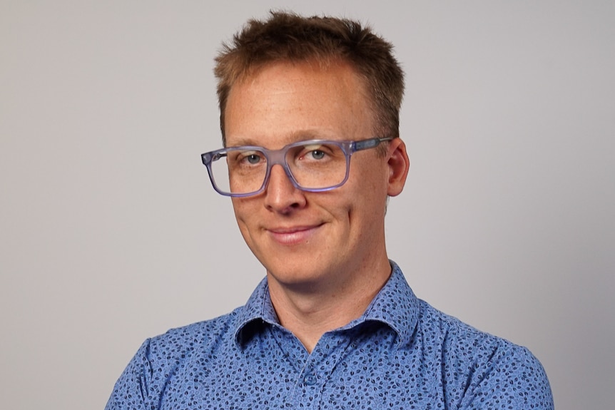 A smiling man with short, dark hair wears glasses and a collared shirt.