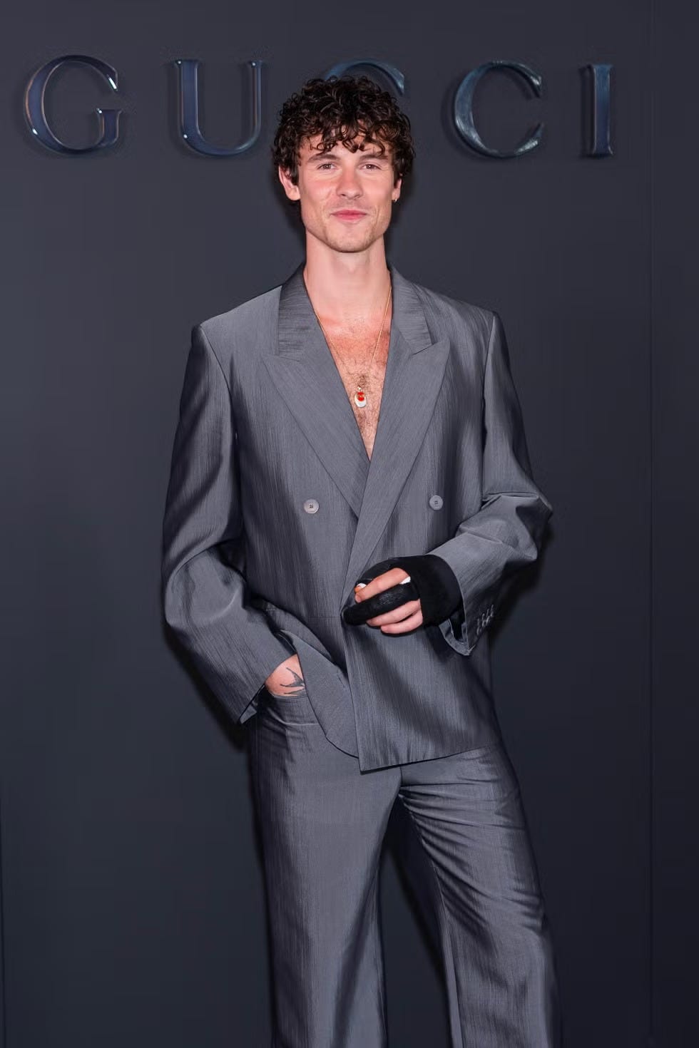 person in a shiny gray suit with a glove, standing against a backdrop with "gucci" letters.