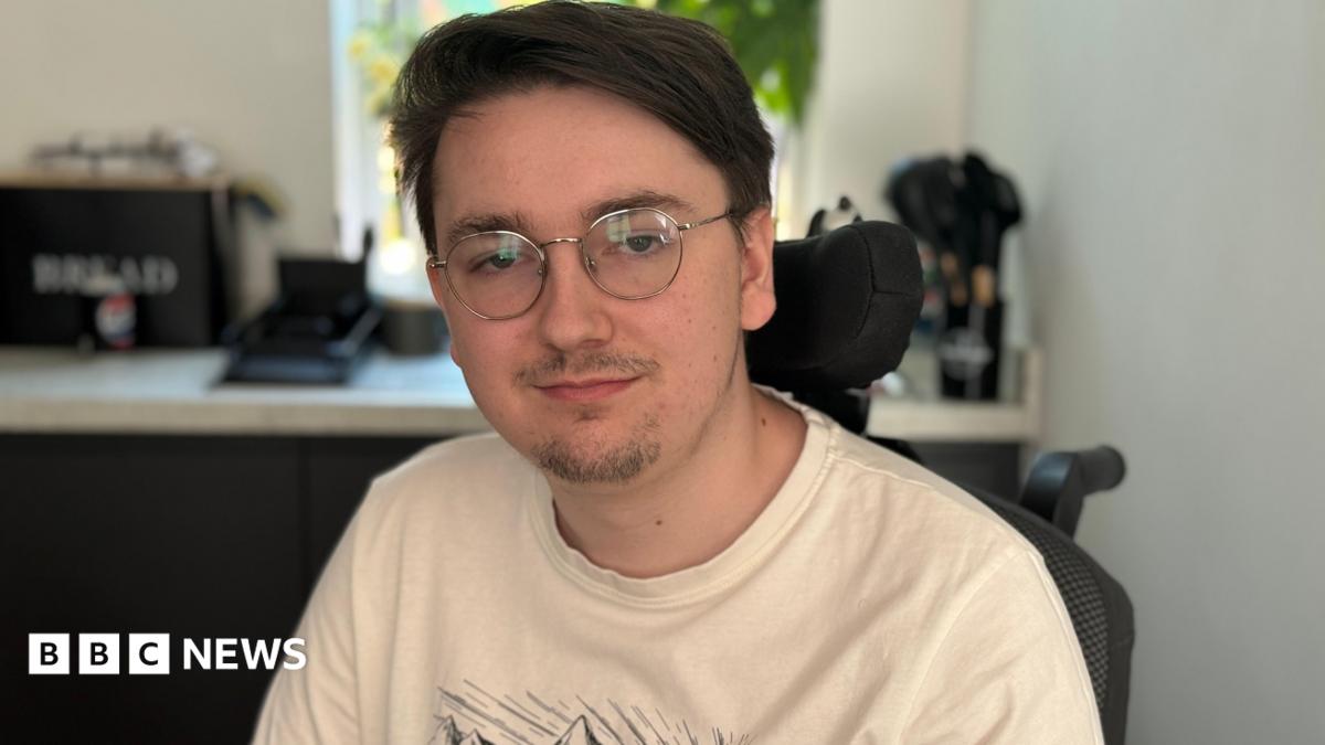 Jack is wearing glasses and a cream T-shirt. He is sitting in his wheelchair in the kitchen
