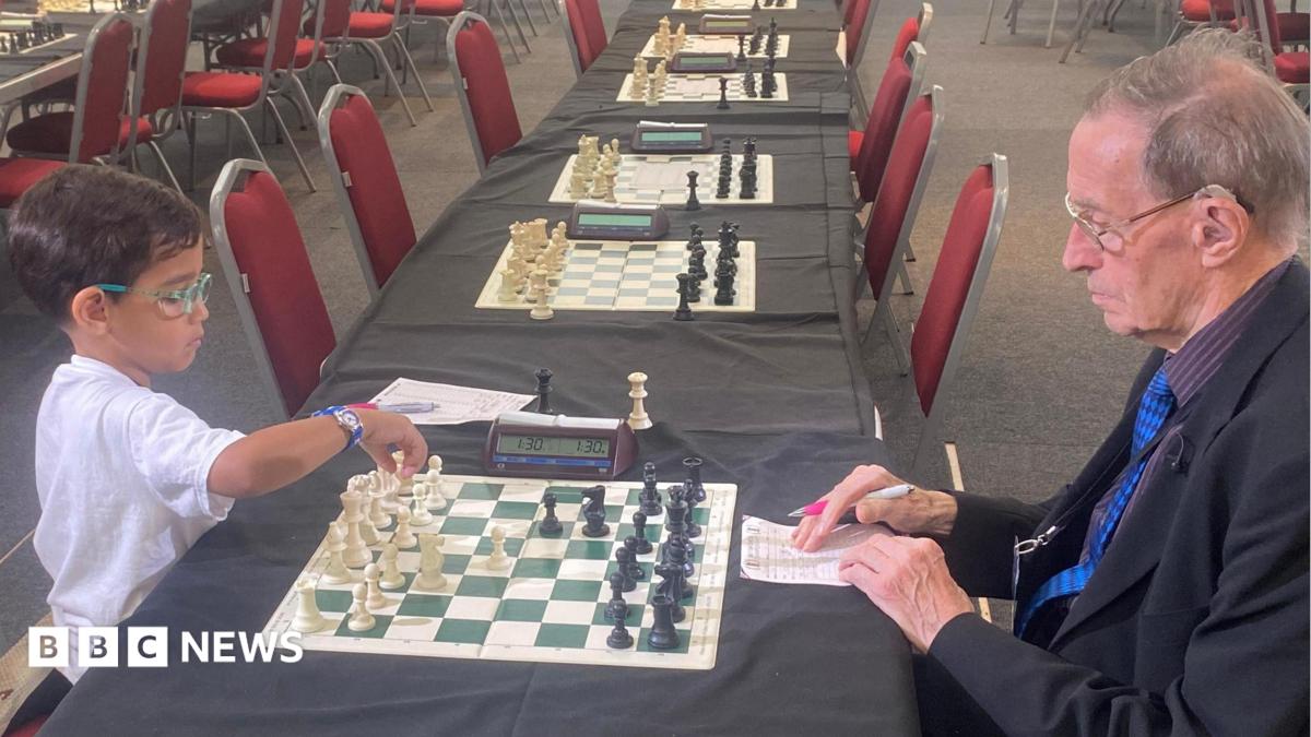 A young boy wearing a white shirt and colourful glasses, with short dark hair sits on a red chair at a table opposite a man with grey hair, wearing a suit and glasses, with a newspaper rolled up in his pocket. They are playing chess. There are many other chess boards and chairs lined up along tables in the room.