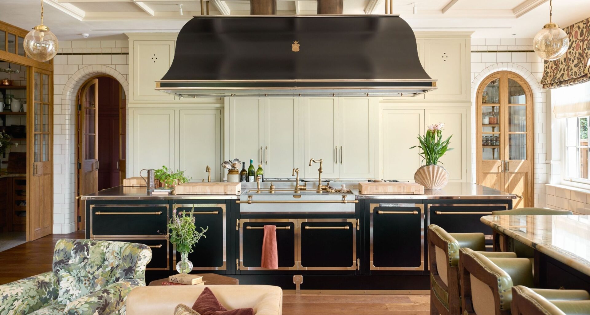 a grand traditional kitchen in Surrey, England with large black and brass island and cook hood, traditional off white cabinetry and arched doors through to pantry spaces