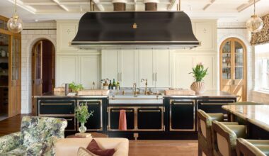 a grand traditional kitchen in Surrey, England with large black and brass island and cook hood, traditional off white cabinetry and arched doors through to pantry spaces