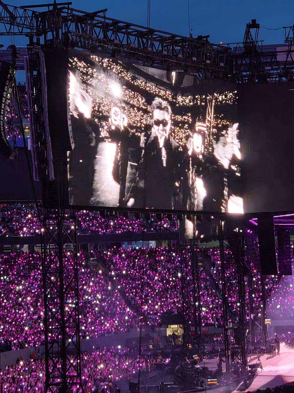 A live concert scene with a large crowd holding purple lights and a performer on a big screen. A live concert scene with a large crowd holding purple lights and a performer on a big screen.