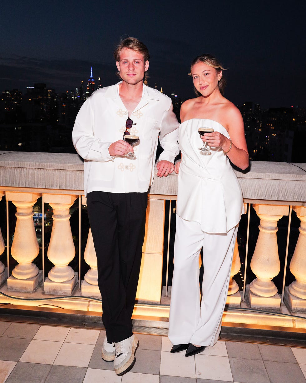 Two individuals holding drinks, standing on a balcony with city lights in the background at night.