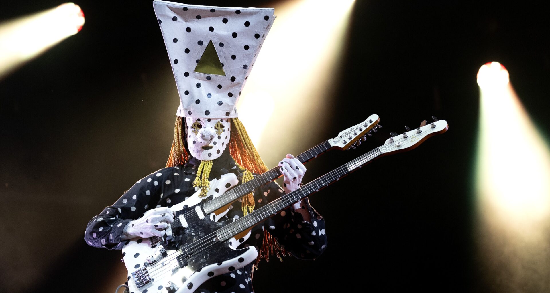 Band member dressed in spotted hat performs under spotlights.