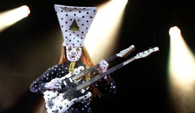 Band member dressed in spotted hat performs under spotlights.