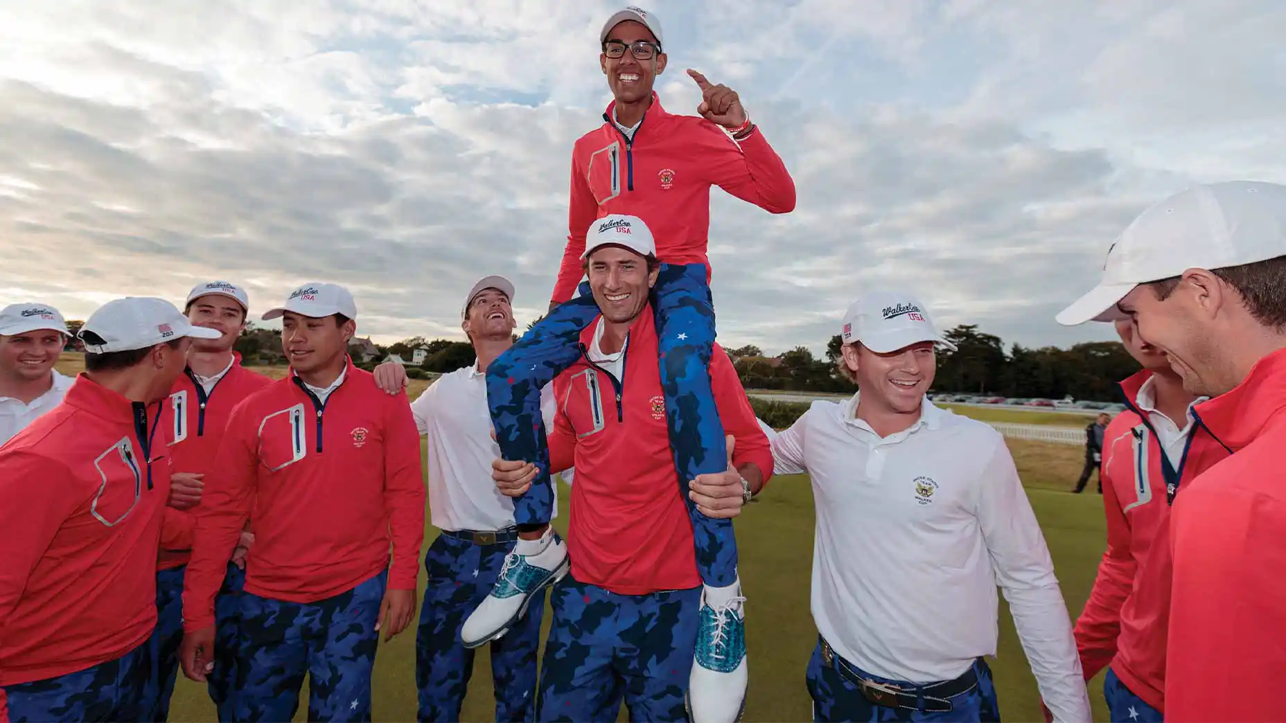 A group of golfers celebrate outdoors; one man in a red jacket is lifted on another’s shoulders while teammates in red and white smile and cheer, standing on a grassy field under a cloudy sky, reminiscent of the camaraderie seen at the Curtis Cup.