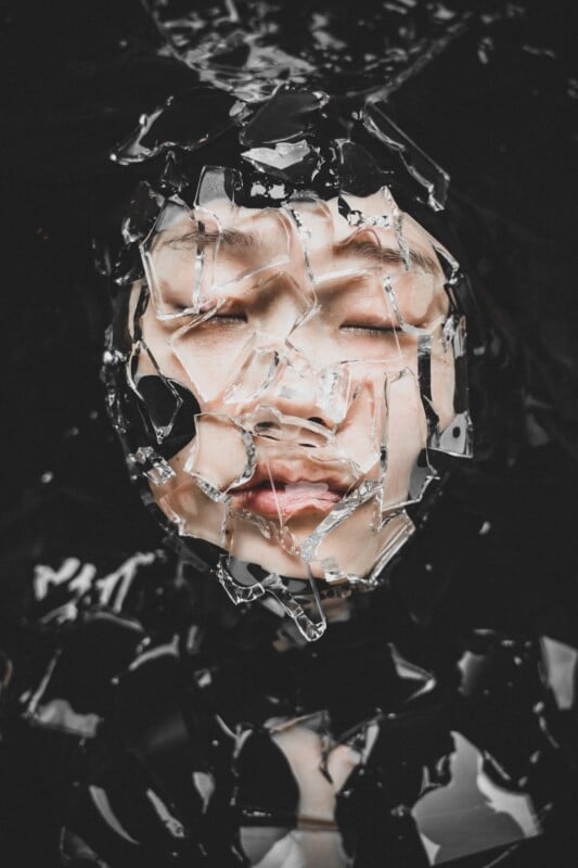 A person's face is partially obscured by shards of broken glass, with their eyes closed against a dark, liquid background.