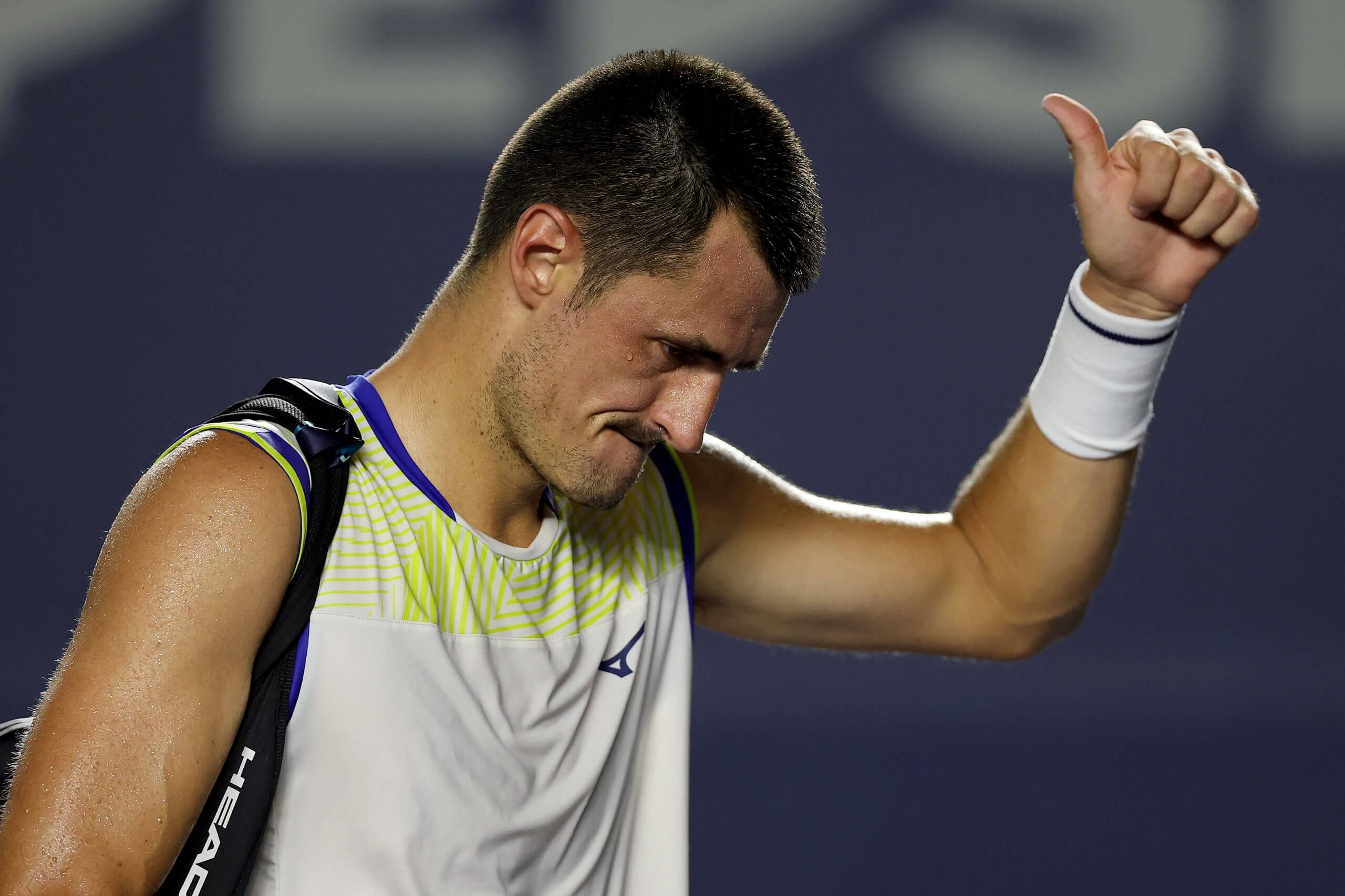 Bernard Tomic raises his left thumb as he walks off a tennis court carrying a bag and wearing a tank top.