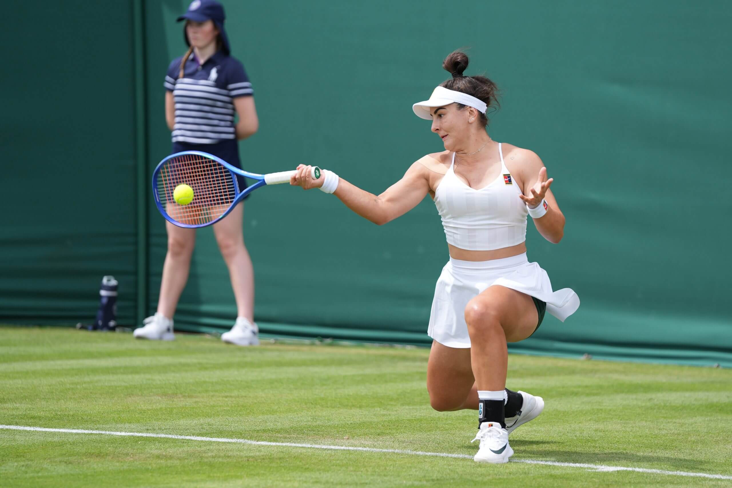 Bianca Andreescu hits a low forehand on a grass tennis court with a ball kid in the backgroubd.