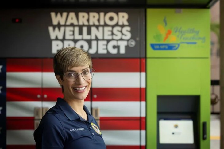 A person stands in front of lockers labeled “Warrior Wellness Project®” and a green unit with an illustration of food and text reading “Healthy Teaching Kitchen.”