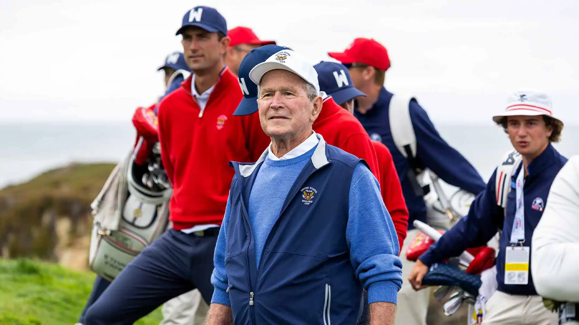 Older man in a blue vest and white cap stands among a group of people in red and blue jackets on a golf course, with golf bags visible in the background—an atmosphere reminiscent of gatherings at Curtis Cup and Walker Cup events, where generous donors and supporters come together to celebrate the spirit of the game.
