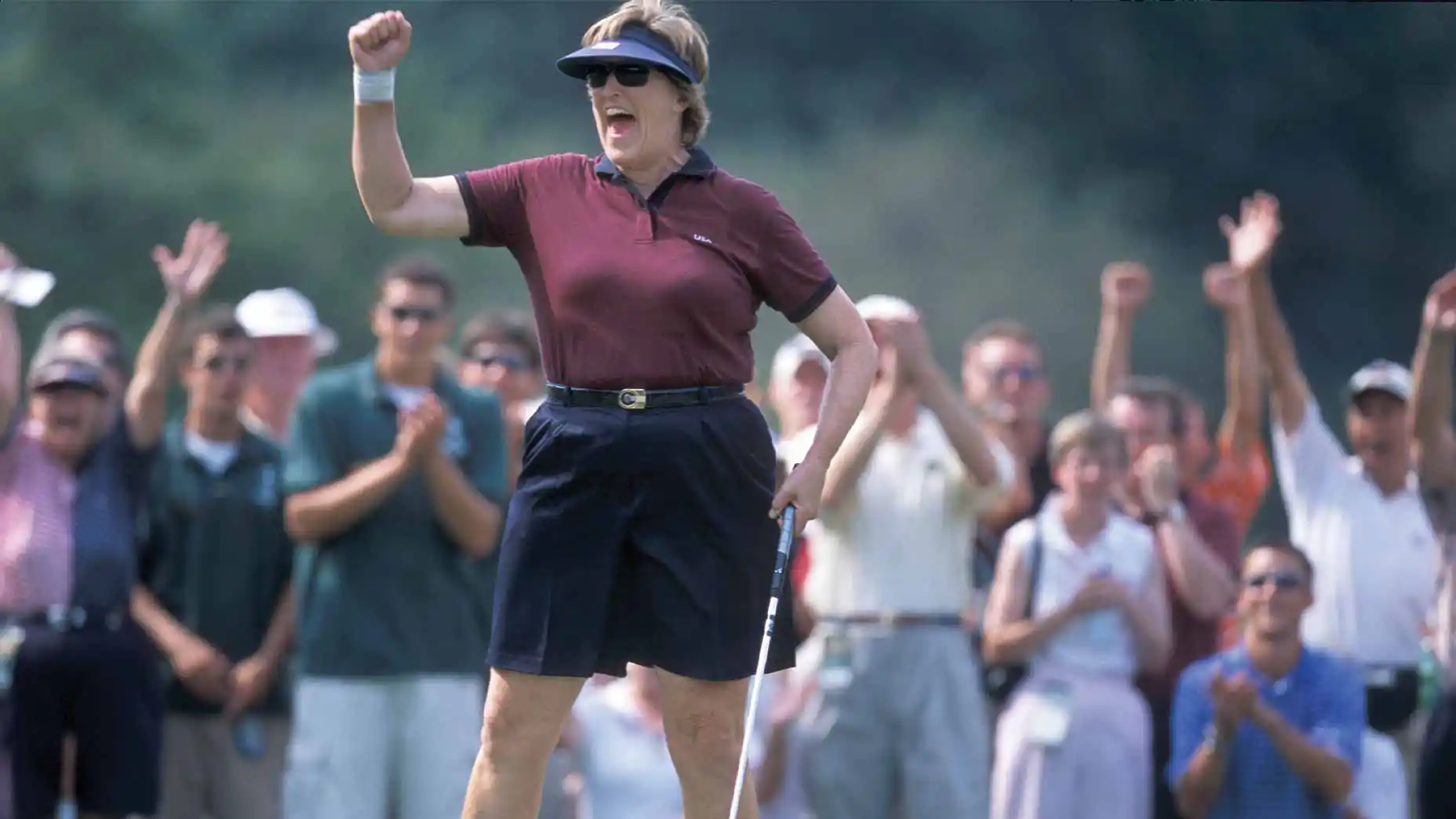 A woman golfer celebrates with a raised fist while holding a club, reminiscent of the spirited moments seen at events like the Curtis Cup. She wears a visor, sunglasses, maroon shirt, and shorts as a crowd in the background—possibly including generous donors—cheers enthusiastically.