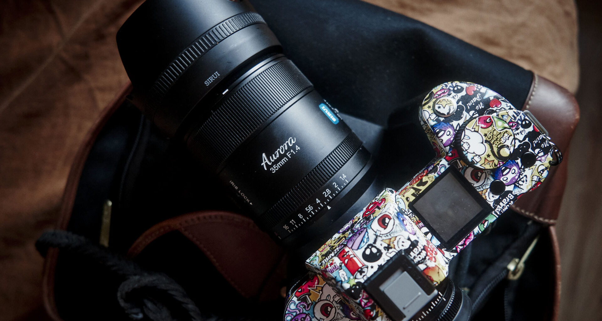 A colorful, sticker-covered camera with a large lens rests on a bag and brown surface.
