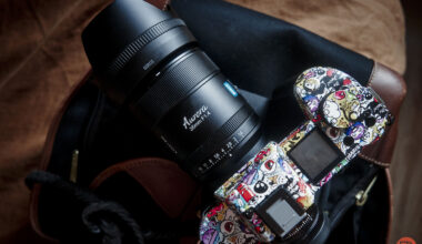 A colorful, sticker-covered camera with a large lens rests on a bag and brown surface.