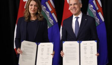 Premier Danielle Smith, a white woman with shoulder-length dark hair, and Prime Minister Mark Carney, a grey-haired white man, each smile and hold documents.