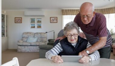 An older gentleman leans over his partner's shoulder as she looks at a tablet device while seated at a table.