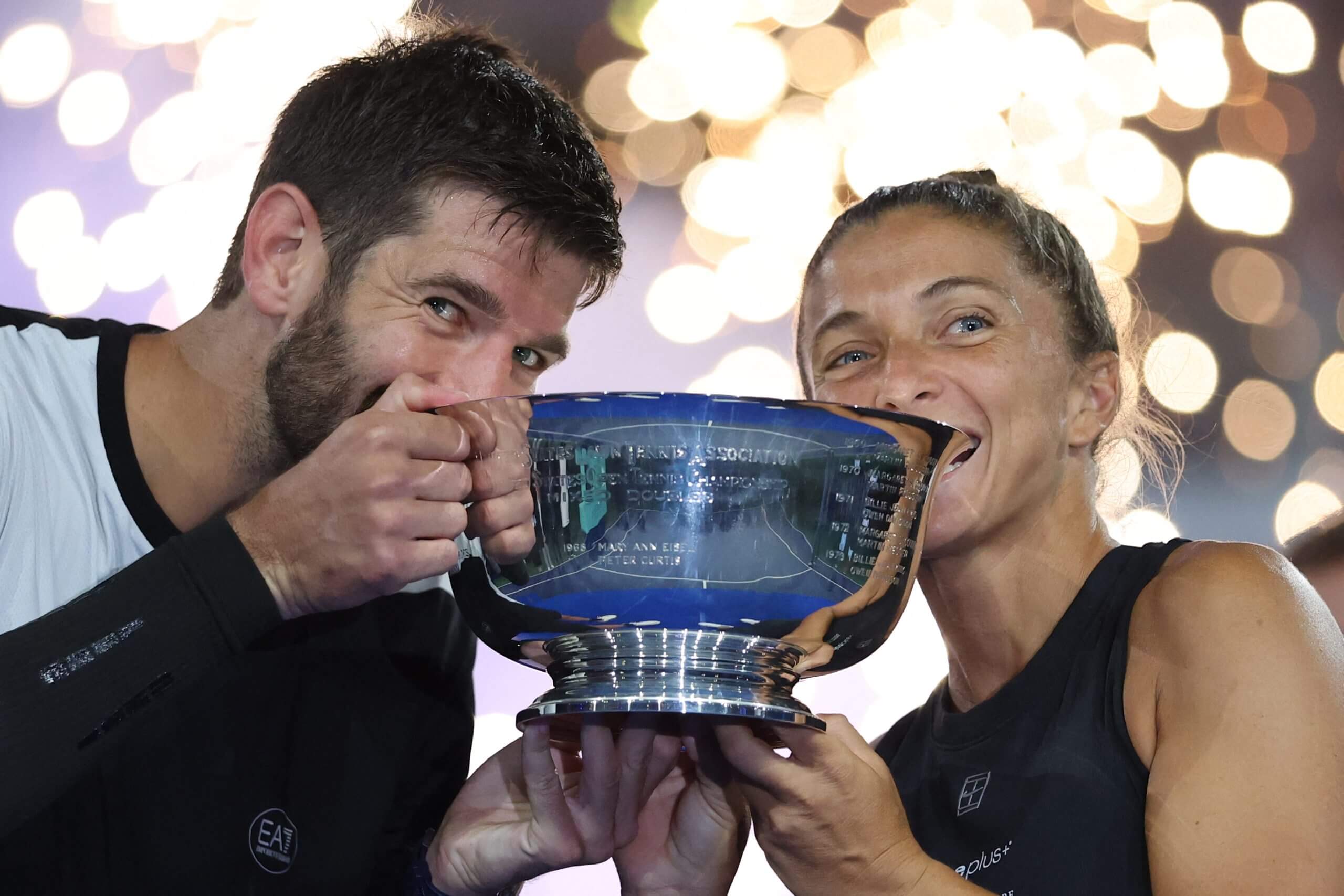 Andrea Vavassori (left) and Sara Errani (right) bite down on a trophy with sparkling lights behind them.