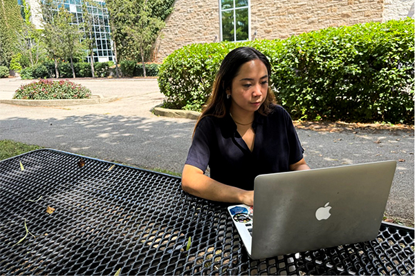 Patricia Mallari working on a laptop at an outdoor table on a university campus.