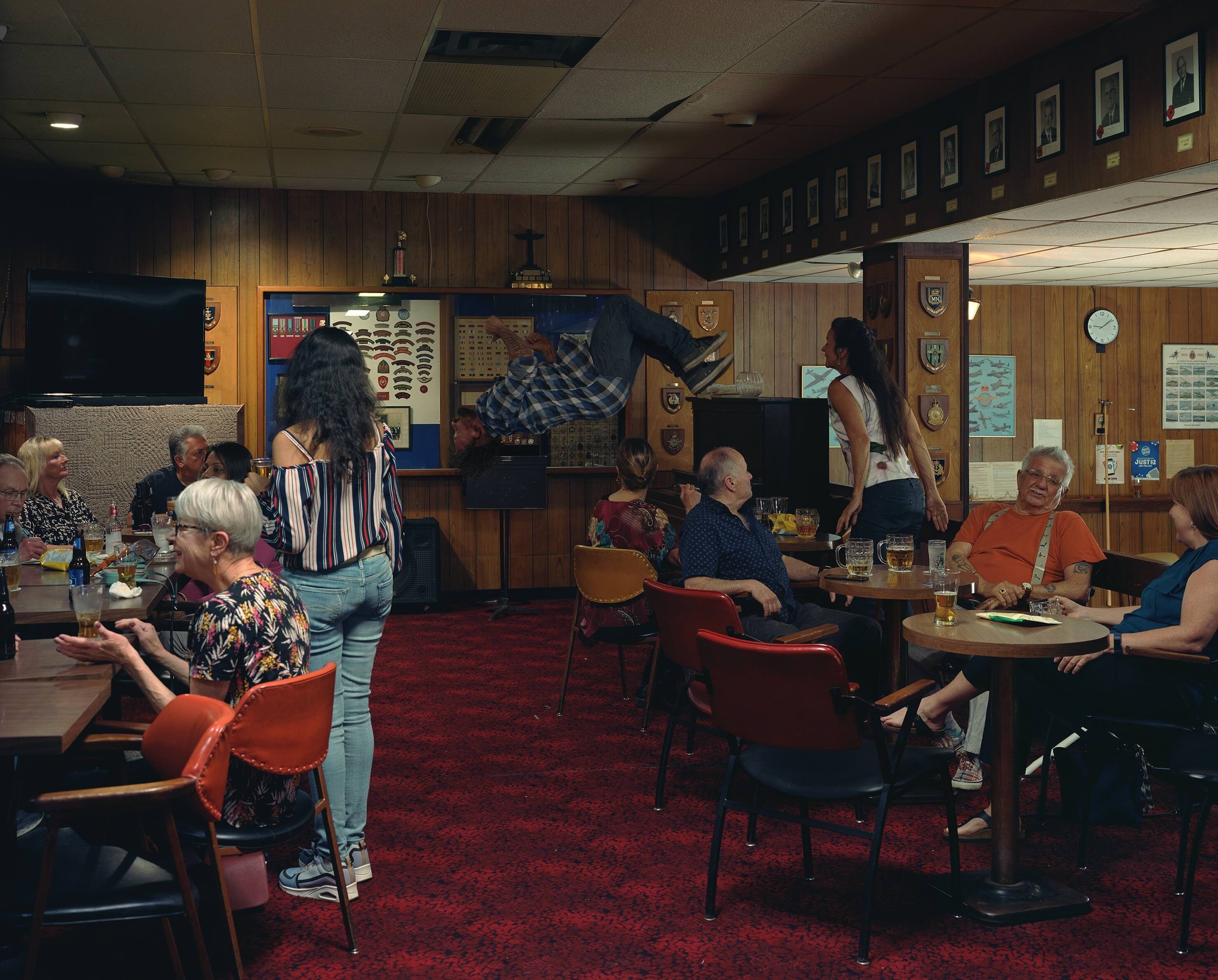 Jeff Wall’s 'In the Legion' is a conceptual photograph that depicts the dimly lit interior of a legion hall where people are drinking beer and socializing. In the background a man is doing a backflip.