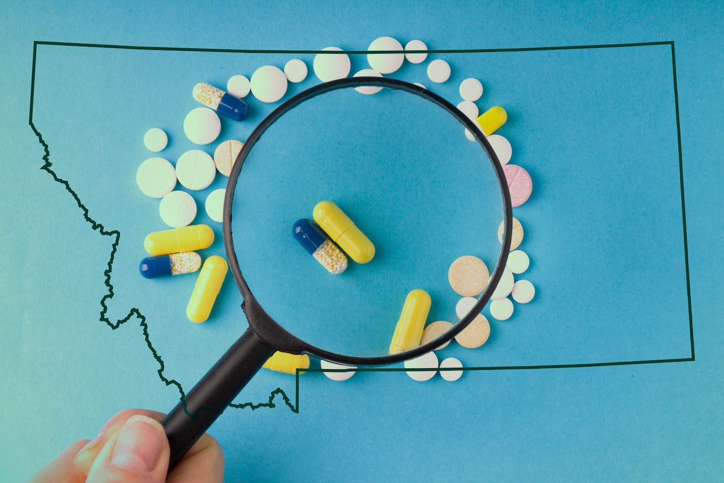 A photo illustration of a hand holding a magnifying glass over a scattering of prescription pills, outlined by the shape of Montana.