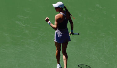 Female tennis player on a green court, seen from behind, wearing a cap and white skirt, holding a racket in her right hand.