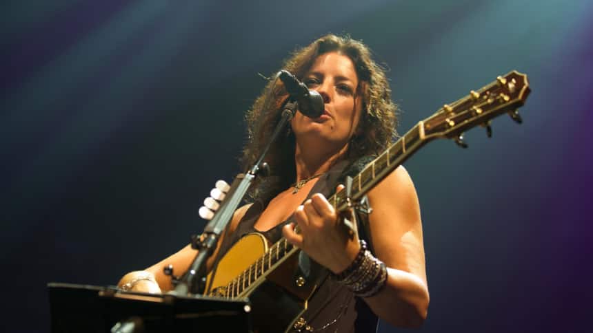 A woman stands playing a guitar and singing into a microphone.